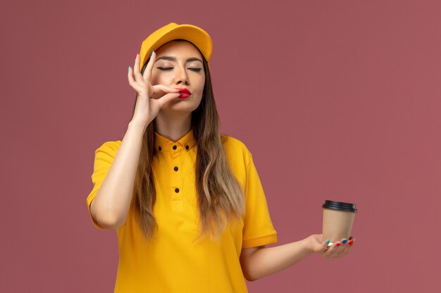 Front view of female courier in yellow uniform and cap holding delivery coffee cup on pink wall