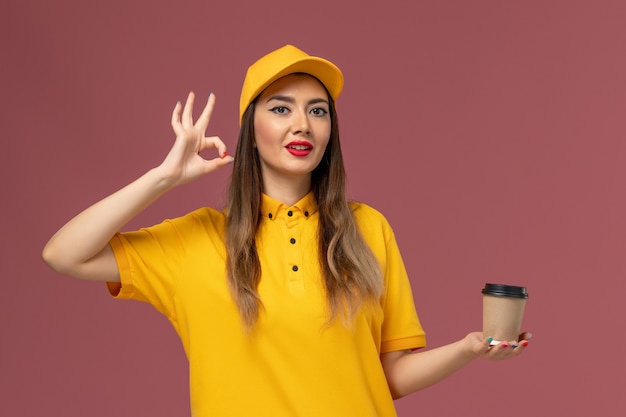 Front view of female courier in yellow uniform and cap holding delivery coffee cup on the pink wall