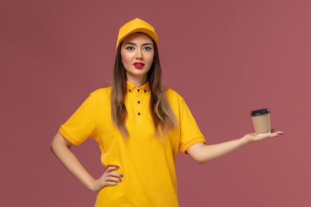 Front view of female courier in yellow uniform and cap holding delivery coffee cup on the pink wall