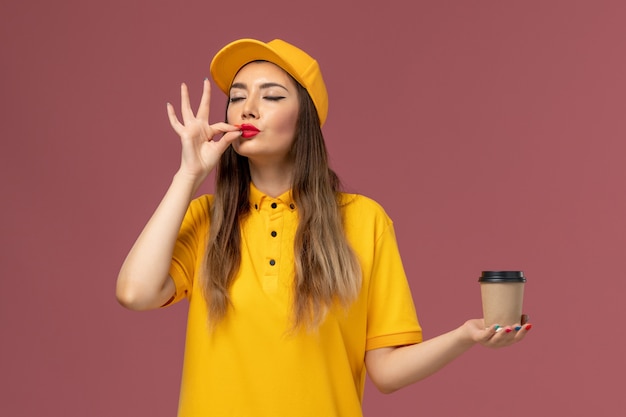 Front view of female courier in yellow uniform and cap holding delivery coffee cup on the pink wall