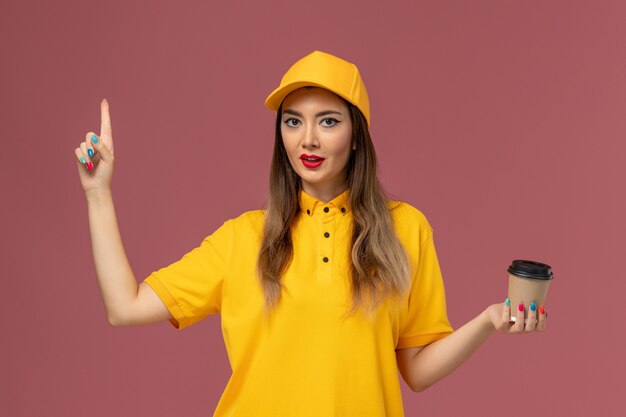Front view of female courier in yellow uniform and cap holding delivery coffee cup on the pink wall