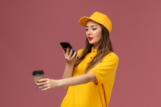 Front view of female courier in yellow uniform and cap holding delivery coffee cup and taking photo of it on pink wall