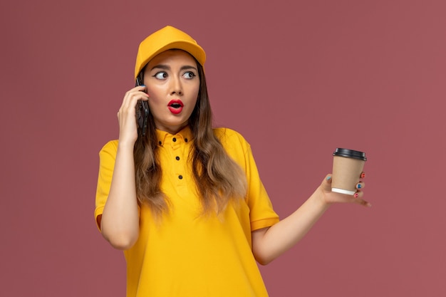 黄色の制服を着た女性の宅配便と配達コーヒーカップを保持し、ピンクの壁に電話で話しているキャップの正面図