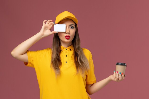 Front view of female courier in yellow uniform and cap holding delivery coffee cup and white card