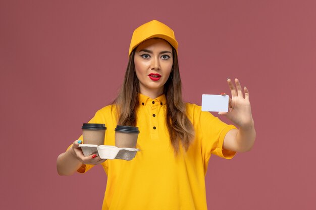 Front view of female courier in yellow uniform and cap holding delivery coffee cups and card on pink wall