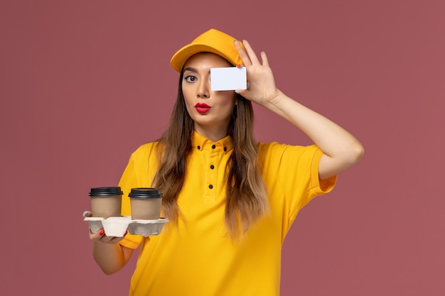 Front view of female courier in yellow uniform and cap holding delivery coffee cups and card on pink wall