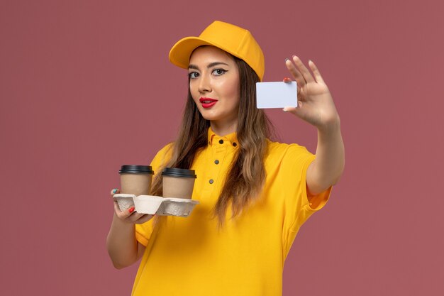 Front view of female courier in yellow uniform and cap holding delivery coffee cups and card on pink wall