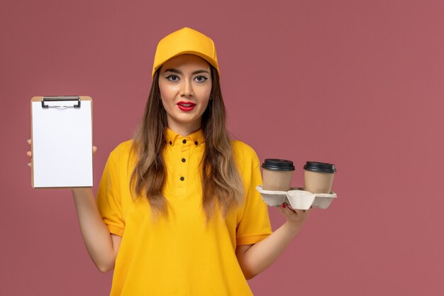 Front view of female courier in yellow uniform and cap holding delivery coffee cups and notepad on light pink wall