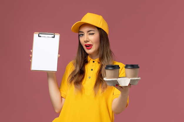 밝은 분홍색 벽에 노란색 유니폼과 모자 배달 커피 컵 메모장을 들고 여성 택배의 전면보기