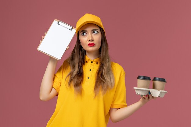 Front view of female courier in yellow uniform and cap holding delivery coffee cups notepad thinking on light pink wall