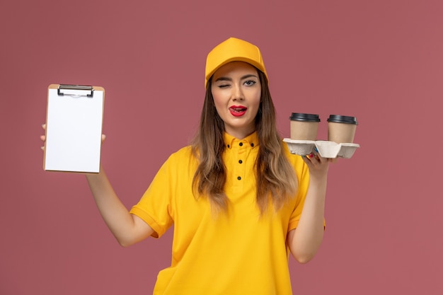 Front view of female courier in yellow uniform and cap holding delivery coffee cups and notepad winking on pink wall