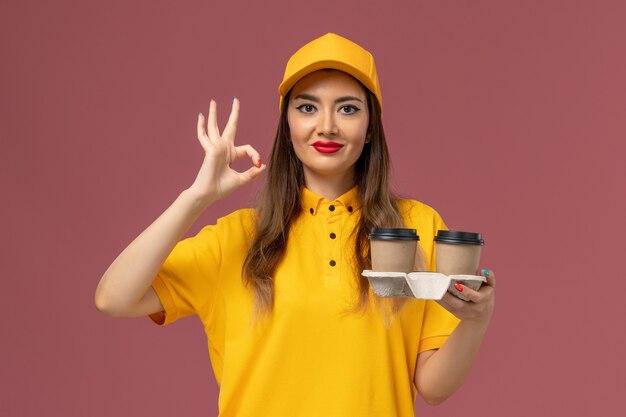 Front view of female courier in yellow uniform and cap holding delivery coffee cups on pink wall