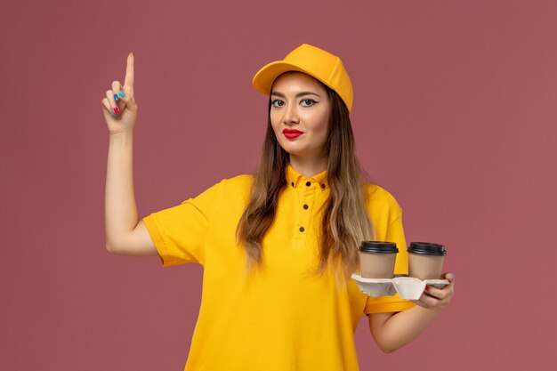 Front view of female courier in yellow uniform and cap holding delivery coffee cups on pink wall