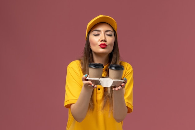 Vista frontale del corriere femminile in uniforme gialla e cappuccio che tiene le tazze di caffè di consegna sulla parete rosa