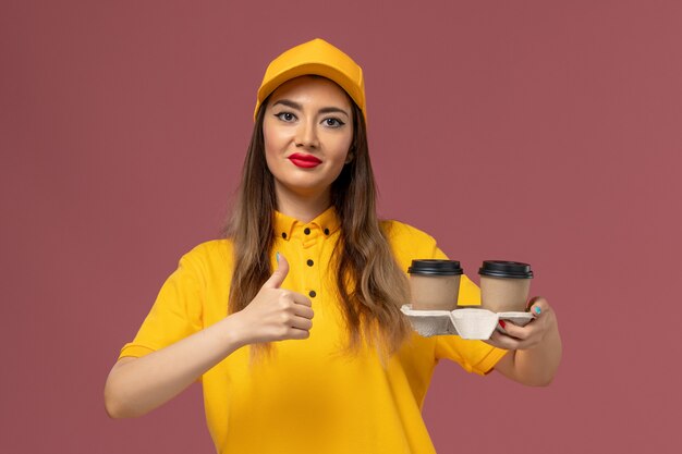 Front view of female courier in yellow uniform and cap holding delivery coffee cups on pink wall