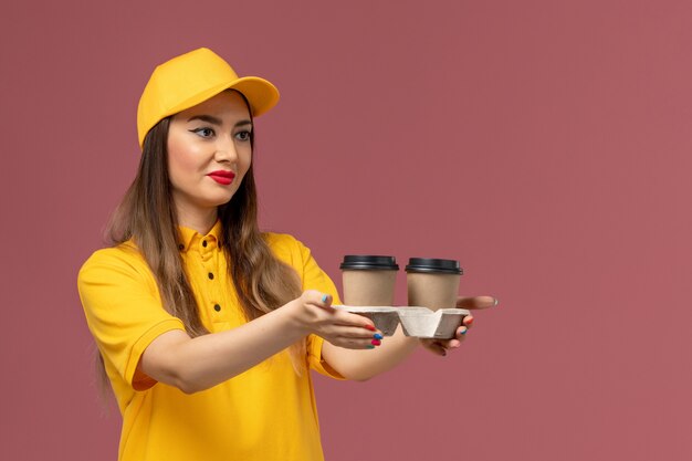 분홍색 벽에 배달 커피 컵을 들고 노란색 유니폼과 모자에 여성 택배의 전면보기