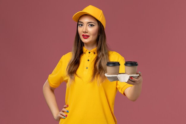 Front view of female courier in yellow uniform and cap holding delivery coffee cups on pink wall