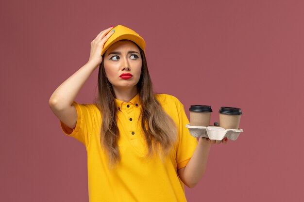 Front view of female courier in yellow uniform and cap holding delivery coffee cups on the pink wall