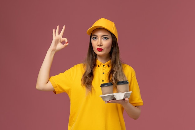 Front view of female courier in yellow uniform and cap holding delivery coffee cups on the pink wall