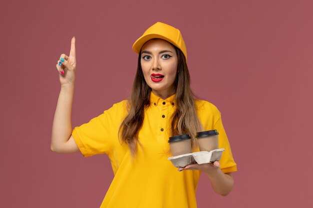 Front view of female courier in yellow uniform and cap holding delivery coffee cups on the pink wall