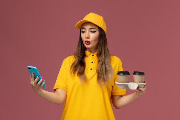 Front view of female courier in yellow uniform and cap holding delivery coffee cups and smartphone on pink wall