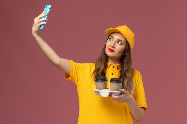 Front view of female courier in yellow uniform and cap holding delivery coffee cups taking photo on pink wall