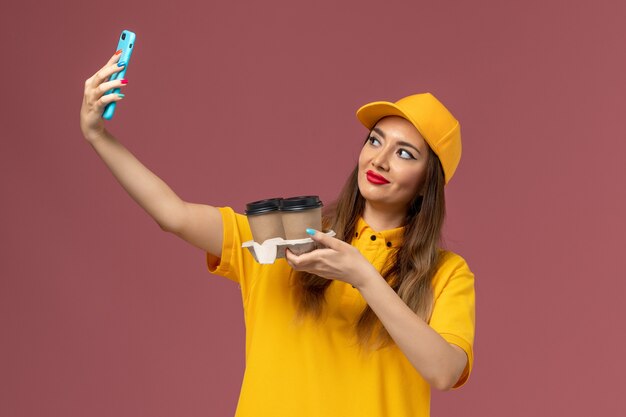 Front view of female courier in yellow uniform and cap holding delivery coffee cups taking a selfie on pink wall