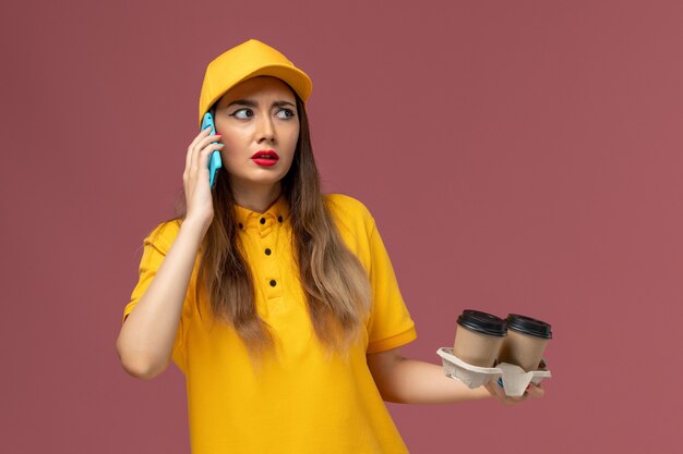 Front view of female courier in yellow uniform and cap holding delivery coffee cups and talking on the phone on the pink wall