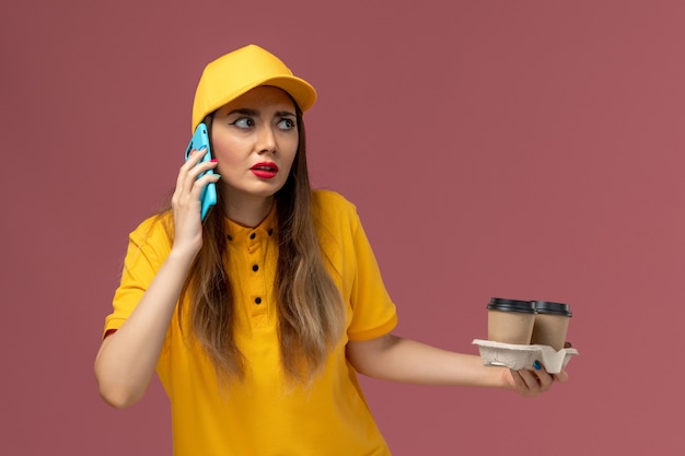 Front view of female courier in yellow uniform and cap holding delivery coffee cups talking on the phone on pink wall