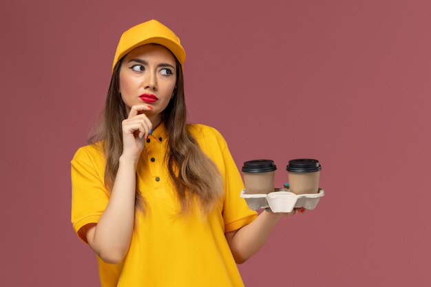 Вид спереди курьера-женщины в желтой униформе и кепке, держащего кофейные чашки с доставкой и думающего о розовой стене