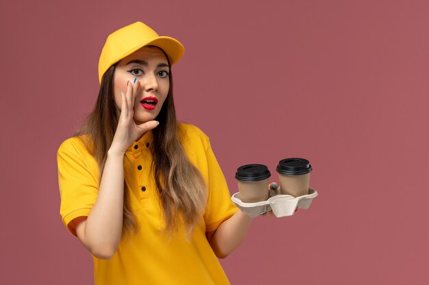 Front view of female courier in yellow uniform and cap holding delivery coffee cups and whispering on the pink wall