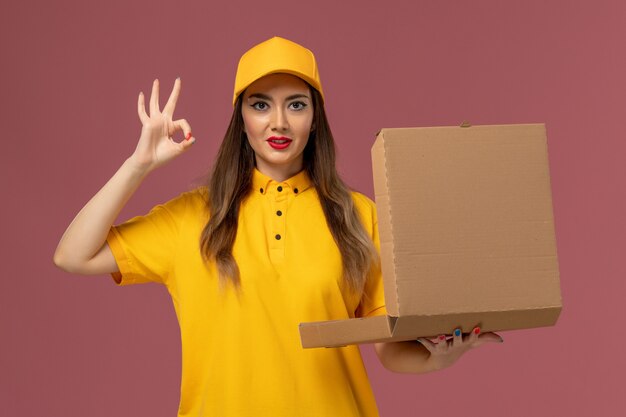 Front view of female courier in yellow uniform and cap holding empty food box on light pink wall