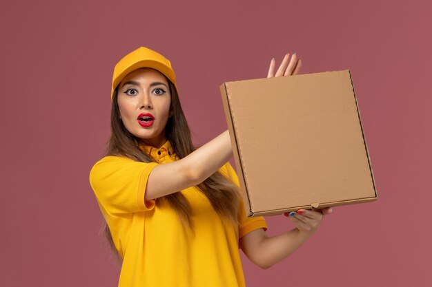 Front view of female courier in yellow uniform and cap holding food box on light-pink wall