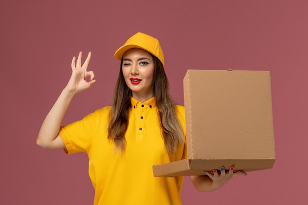 Вид спереди курьера в желтой форме и кепке, держащего коробку с едой на светло-розовой стене