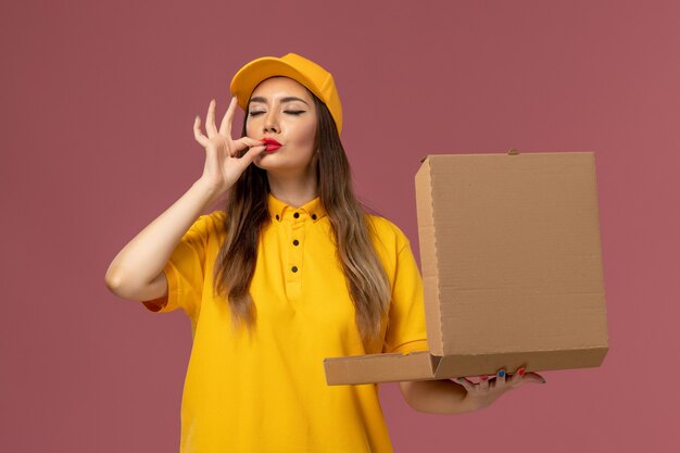 Front view of female courier in yellow uniform and cap holding food box on light pink wall