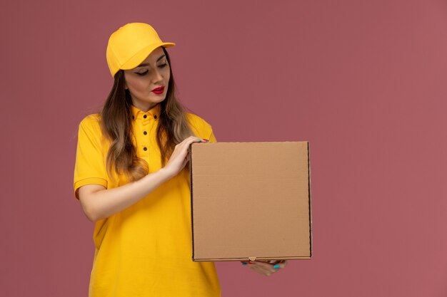 Front view of female courier in yellow uniform and cap holding food box on the light-pink wall