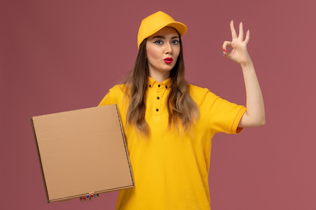 Front view of female courier in yellow uniform and cap holding food box on the light-pink wall