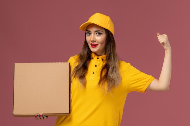 Front view of female courier in yellow uniform and cap holding food box on the light-pink wall