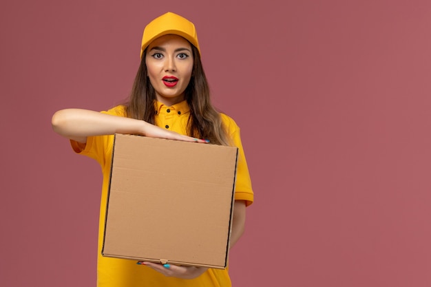 Front view of female courier in yellow uniform and cap holding food box on the light-pink wall