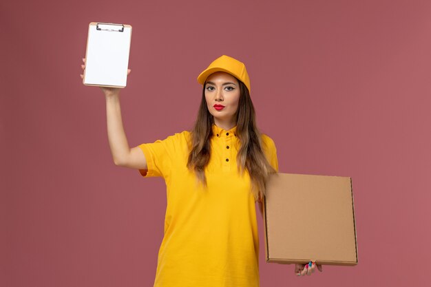 Front view of female courier in yellow uniform and cap holding food box and notepad on the light pink wall