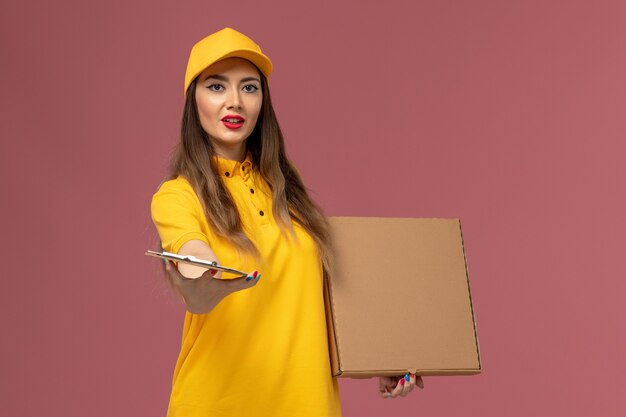 Вид спереди курьера в желтой форме и кепке, держащего коробку с едой и блокнот на розовой стене