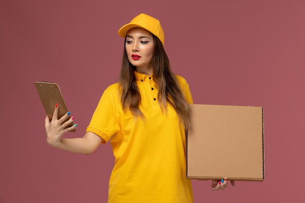 Front view of female courier in yellow uniform and cap holding food box and notepad on pink wall