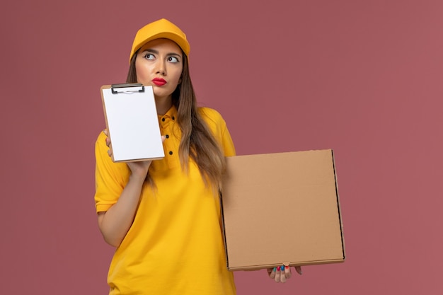 Front view of female courier in yellow uniform and cap holding food box and notepad thinking on pink wall