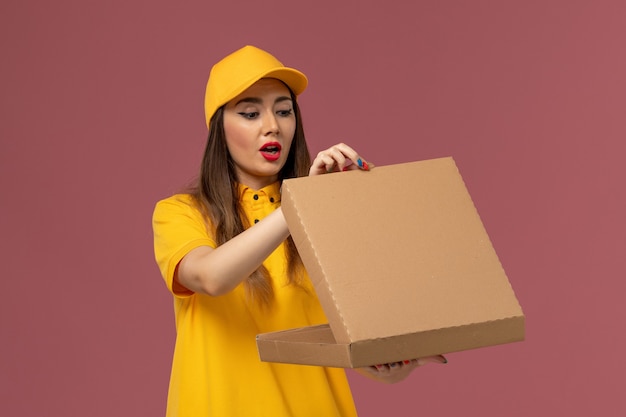 Front view of female courier in yellow uniform and cap holding food box opening it on light pink wall