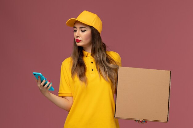 Вид спереди курьера в желтой форме и кепке, держащего коробку для еды и телефон на светло-розовой стене