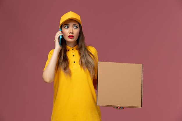 Front view of female courier in yellow uniform and cap holding food box and talking on the phone on light pink wall