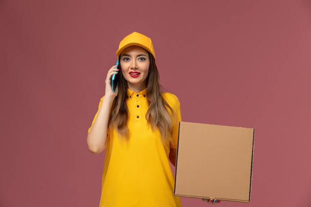 黄色の制服とキャップがフードボックスを保持し、淡いピンクの壁に電話で話している女性の宅配便の正面図