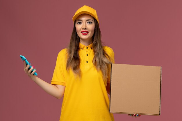 Front view of female courier in yellow uniform and cap holding food box and work phone on light pink wall