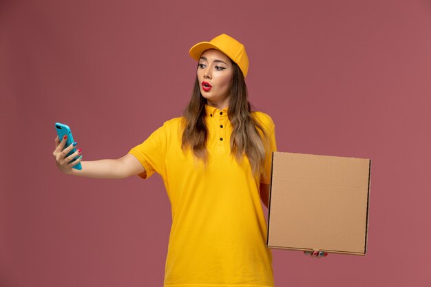 Вид спереди курьера-женщины в желтой форме и кепке, держащей коробку для еды и рабочий телефон на светло-розовой стене