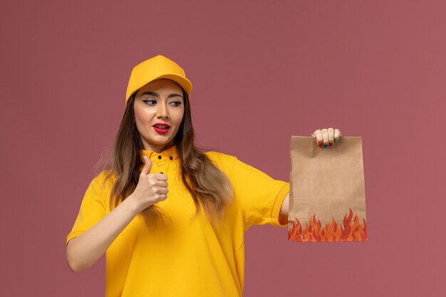 Front view of female courier in yellow uniform and cap holding food package on light-pink wall